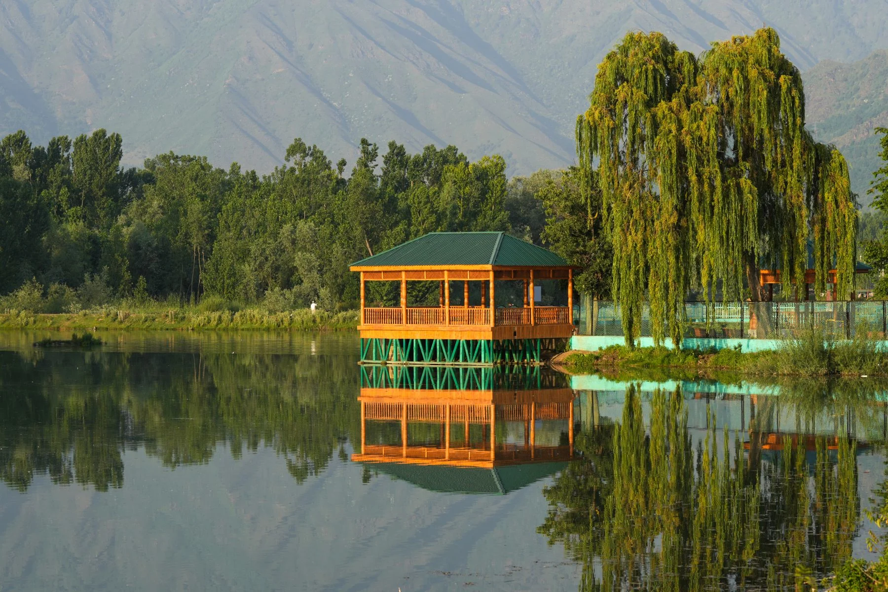 A wooden pavilion reflected in Dal Lake, with willow trees and the mountains of Kashmir beyond